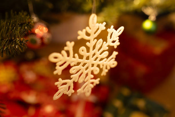 A delicate white snowflake ornament hanging on a beautifully decorated Christmas tree, illuminated by warm festive lights. Blurred wrapped gifts in the background enhance the holiday atmosphere, making it a perfect representation of the magic and joy of the Christmas season.