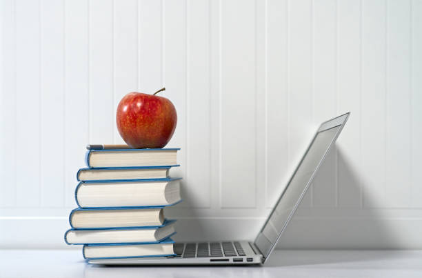Open slim laptop with a stack of blue books on it and a red apple and pencil on top, a white desk, white wainscoting.
