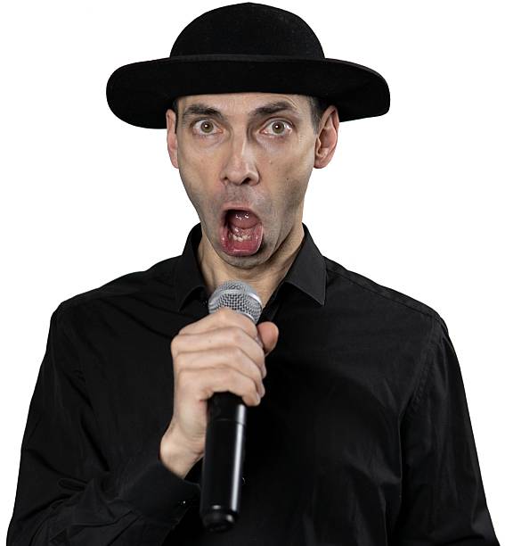 A man in a black shirt and hat sings into a microphone with a funny expression on his face. His animated face and dynamic pose convey the energy and humor of the moment, making it a lively and exciting scene. The background is transparent