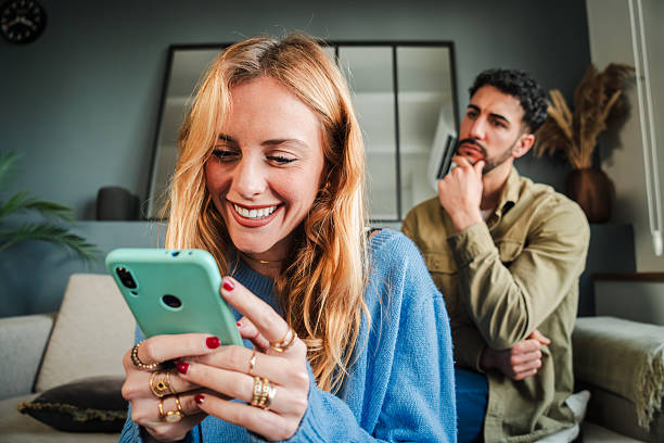 istock Woman using her mobile phone to cheat on her husband, while he spies on her over her shoulder and reads her conversation. Angry and displeased boyfriend observes his girlfriend while she ignores him 2205867560
