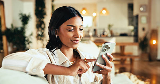 istock Home, smartphone and woman scroll on app, reading blog or post comment on online forum for chat. Living room, mobile and happy Indian girl relax on social media, notification or play game on internet 2205872369