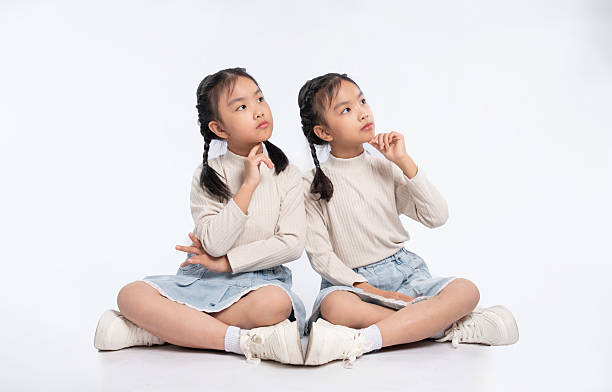 istock Twins pondering, seated cross-legged, deep in thought, with matching outfits, against a white backdrop. 2205873525