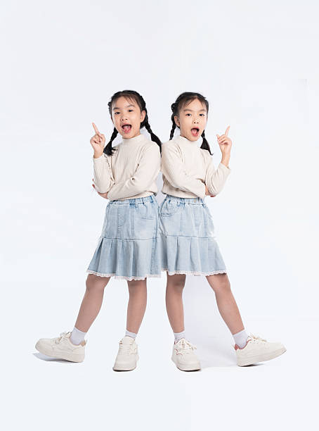 istock Identical twins strike a playful pose, pointing upwards in matching outfits, on a bright, neutral backdrop. 2205876774