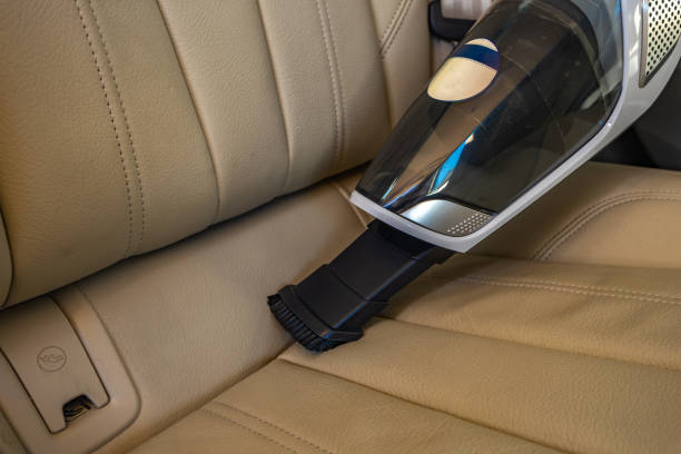 Person using a vacuum cleaner to clean the interior of a car, focusing on seat and central console areas. Depicts detailed cleaning and maintenance of the vehicle for hygiene and cleanliness.