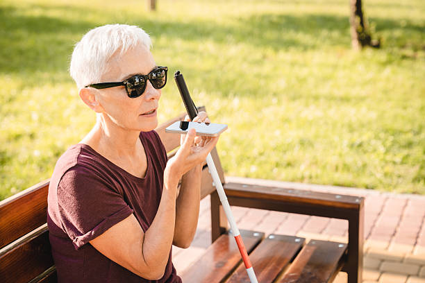 An elderly blind woman sits on a bench in the park and talks on a smartphone. A visually impaired elderly woman wearing sunglasses communicates using a smartphone, seated on a park bench.