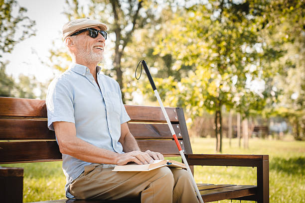 istock Side view of senior vision-impaired blind man sits on a park bench under sunlight, reading a book in braille. He is accompanied by a white cane and surrounded by lush green foliage in nature park 2205881614
