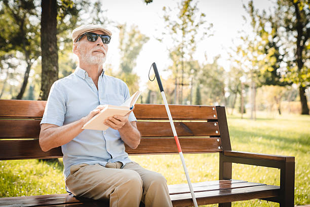 istock A senior vision-impaired blind man sits on a park bench under sunlight, reading a book in braille. He is accompanied by a white cane and surrounded by lush green foliage in nature park copy space 2205881741