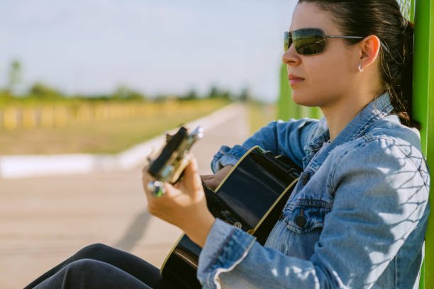A young woman wearing a denim jacket and sunglasses enjoys a peaceful moment while playing the guitar. Sitting outdoors, leaning against a green fence, she conveys a sense of tranquility and connection with music. The natural light and open surroundings create a relaxing atmosphere, perfect for illustrating concepts of inspiration, creativity, and introspective moments.