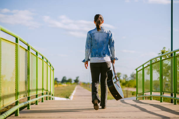Woman walking with her guitar on a sunny day, dressed in a jean jacket and black pants