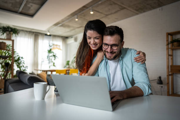 istock Young couple smiling and browsing internet on laptop at home 2205891597
