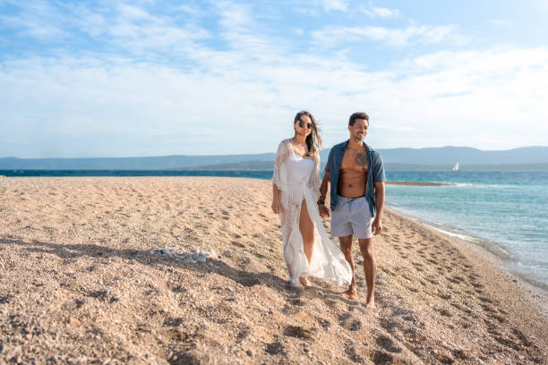 A cheerful mid-adult couple, dressed in casual summer attire, walks barefoot on a sunny beach with sparkling ocean waves and blue skies.
