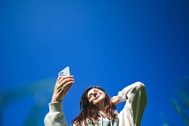 istock Young woman taking a selfie with a smartphone against blue sky 2205896267