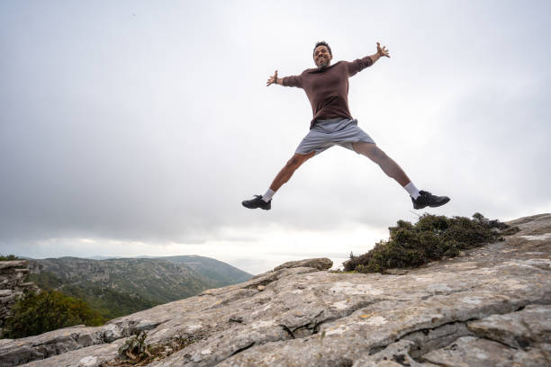 Wide-angle photograph showing mid-adult Black man performing energetic jump with outstretched arms on rocky mountain summit, overlooking dramatic mountainous landscape beneath overcast sky.