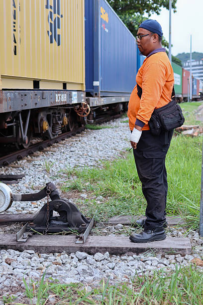 Perak, Malaysia - June 22, 2024 : A railway worker in an orange shirt and black pants stands beside cargo containers on a train at an inland port, highlighting logistics and transportation operations. Ideal for showcasing industrial and railway themes.