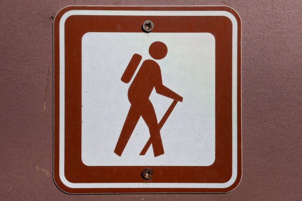 Brown and white sign displays a silhouette of a hiker with a backpack and walking stick, indicating a designated hiking trail. The sign is simple and universally recognizable, often found in national parks, forests, or nature reserves to mark trails for outdoor enthusiasts. This image is ideal for themes related to hiking, outdoor adventures, or trail navigation.