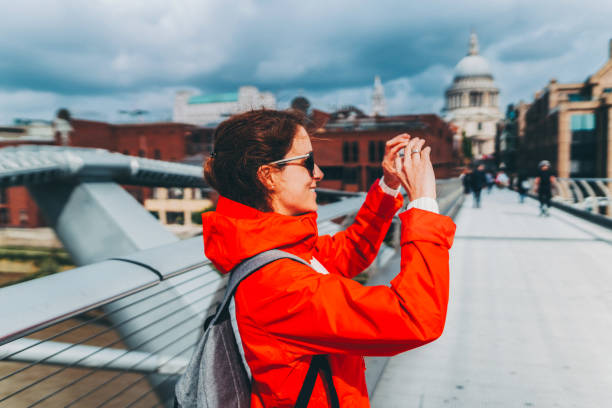 Smiling caucasian young adult female explorer walking in front of an important landmark in London. She is visiting London alone. She look happy to be exploring the city. The woman is looking around as she is walking by the river Thames.She has a backpack on her back and a cool camera around her neck.
