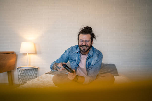 istock Happy man playing videogames sitting on bed in modern apartment 2205928988