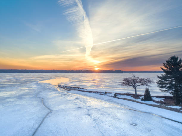 A frozen lake stretches under a vibrant sky, with the sun setting on the horizon, casting warm colors across the ice. Trees frame the scene on one side, adding contrast to the bright reflections. Detroit Point, Higgins Lake, Michigan. 3/13/25