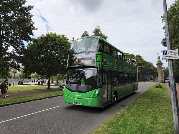 Edinburgh, Scotland, UK - August 3, 2024: Double decker city tour bus, Edinburgh. Edinburgh during the Edinburgh Fringe Festival. There are people walking or relaxing. At the background on the right, one of the garden gates of the Palace of Holyroodhouse and on the left behind the trees Scottish Parliament Building