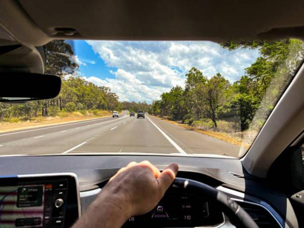 istock A wide-angle view through a car’s windscreen reveals a highway through a forested area, framed by the interior, steering wheel, and driver’s hands under a clear blue sky with fluffy clouds 2205944926