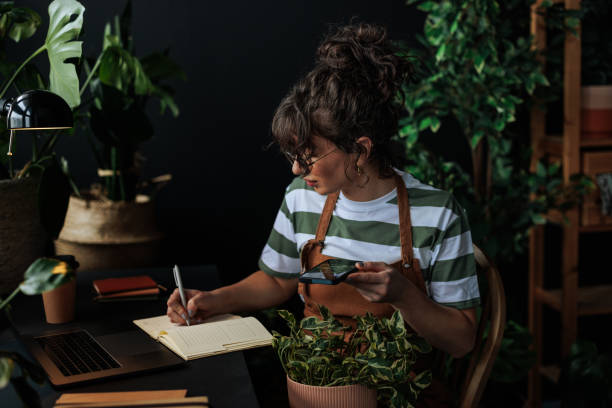 Young woman botanist writing notes and using smart phone in her plant shop