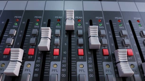 Close-up of an audio mixing console with white faders, LED indicators, and various control buttons for adjusting sound levels.
