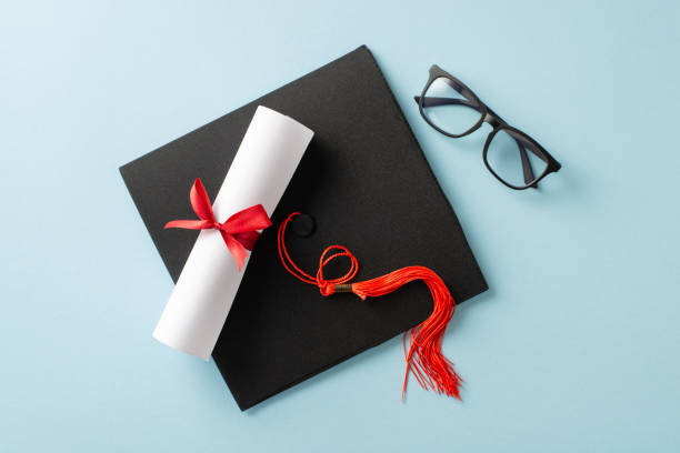 istock Graduation accessories including a diploma with red ribbon and a cap laying on a surface 2205956993