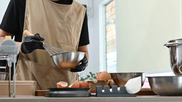 istock A man is making a cake in a kitchen. He is wearing apron and gloves. There are several bowls and spoons on the counter. 2205977099