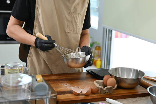 istock A man is making a cake in a kitchen. He is wearing apron and gloves. There are several bowls and spoons on the counter. 2205977113