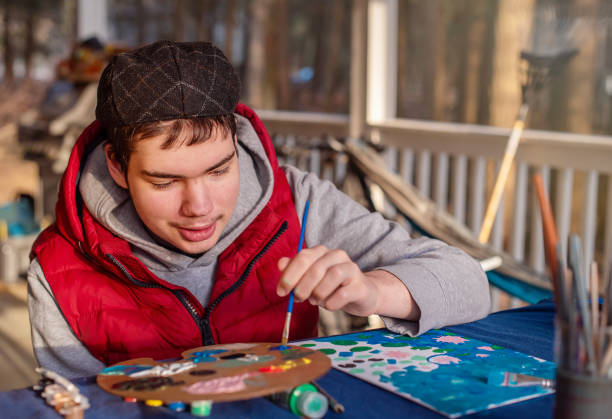 A 19-year-old autistic young man paints a picture as a Mother's Day gift. He wears a brown cap, red vest, and gray hoodie while focusing intently on his artwork. The painting session takes place on the porch of their Pennsylvania home during spring. Art supplies including a paint palette and brushes are arranged on the blue table in front of him.