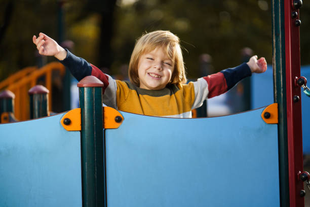 An Asian boy in a navy puffer jacket plays on top of a wooden playground fort, pointing forward with excitement.