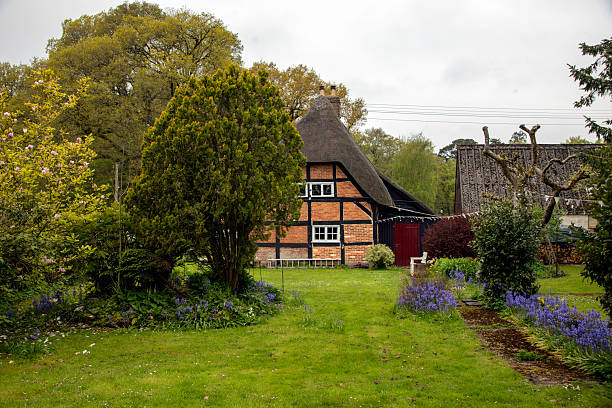 A charming half-timbered thatched cottage is nestled within a lush spring garden in the English countryside. The classic architecture, with its exposed wooden beams and steep thatched roof, reflects traditional British rural design. Surrounded by blooming flowers, manicured grass, and mature trees, the scene evokes a tranquil and timeless atmosphere. Purple wildflowers and a stone path lead the viewer’s eye toward the cozy dwelling, while overcast skies enhance the green tones and peaceful mood. The cottage, flanked by other rustic buildings, embodies the idyllic beauty of rural living in England.