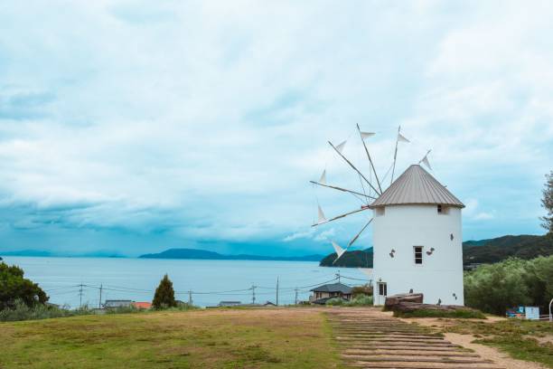 A picturesque view of a white windmill by the sea under a cloudy sky in Shodoshima, Japan.