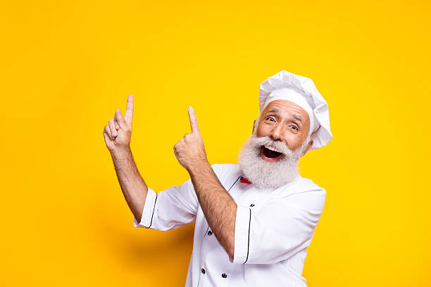 istock Happy senior chef enthusiastically pointing, wearing uniform on yellow background 2206003445