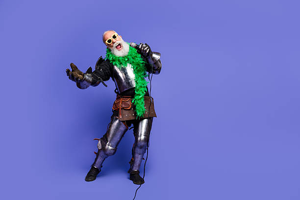 An elderly bearded man wearing a medieval knight costume enthusiastically sings into a microphone against a solid purple background.
