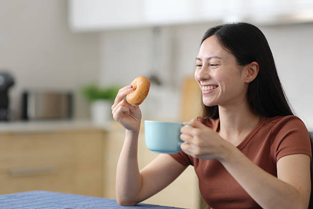 istock Happy asian woman eating doughnut drinking coffee in a kitchen 2206003937