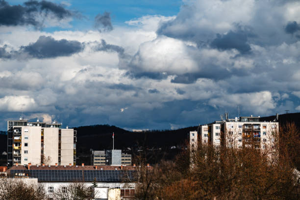 A cityscape featuring multiple residential apartment buildings set against a backdrop of dramatic clouds and rolling hills. The mix of modern infrastructure, solar panels, and natural elements highlights urban development and environmental consciousness. The contrast between the illuminated facades and the dark clouds adds depth and intensity to the composition.