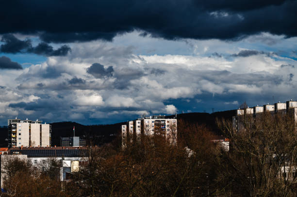 A cityscape featuring multiple residential apartment buildings set against a backdrop of dramatic clouds and rolling hills. The mix of modern infrastructure, solar panels, and natural elements highlights urban development and environmental consciousness. The contrast between the illuminated facades and the dark clouds adds depth and intensity to the composition.
