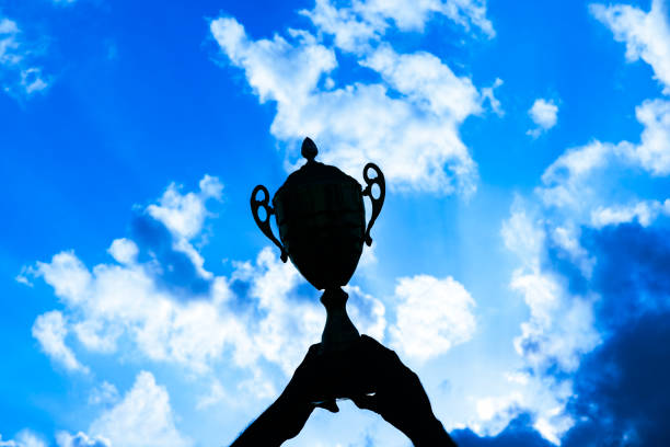 Multiple hands reaching for a silhouetted trophy against a radiant sunset, depicting a team's collective success.