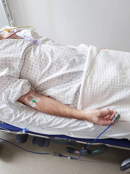 istock IV drip, cannula or branula and a pulse oximeter connected to a Caucasian man's arm. Lying in hospital bed, medical patient gown, close up shot, unrecognizable, clean white bedding 2206023716