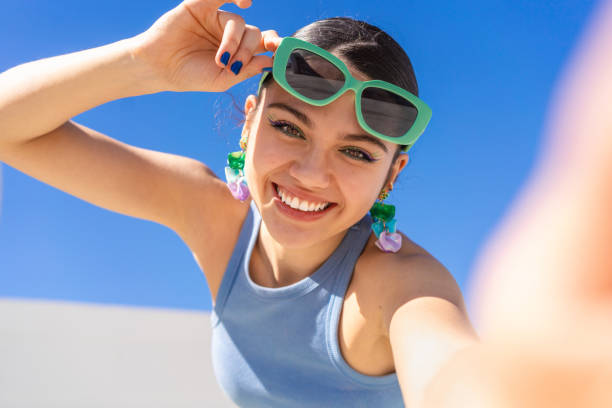 Smiling woman capturing selfie, adjusting stylish sunglasses with bright blue sky backdrop