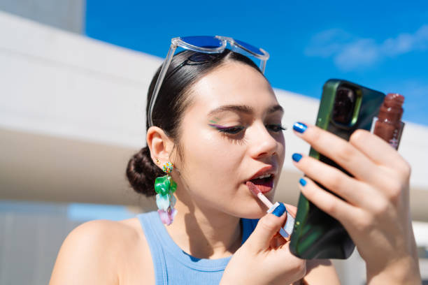 Young woman is applying lip gloss while using her smartphone as a mirror, showcasing modern beauty routines