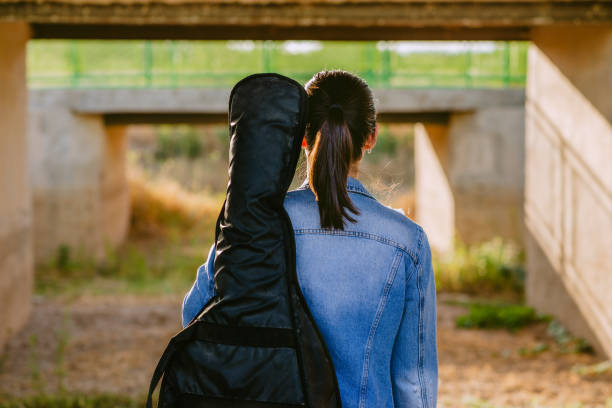 A woman seen from behind, wearing a denim jacket and carrying a guitar case on her shoulder while walking under a bridge. The image evokes a sense of journey, musical inspiration, and exploration. Ideal for projects related to music, creativity, and the nomadic lifestyle.
