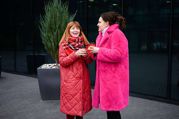 istock Two attractive women in winter coats talking and drinking coffee standing outside during winter walk along city street, smiling, talking, laughing. Women on city street drinking hot drink.Coffee break 2206057235