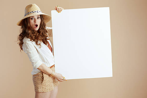 Beach vacation. surprised stylish female in white blouse and shorts isolated on beige background with summer hat showing blank board.