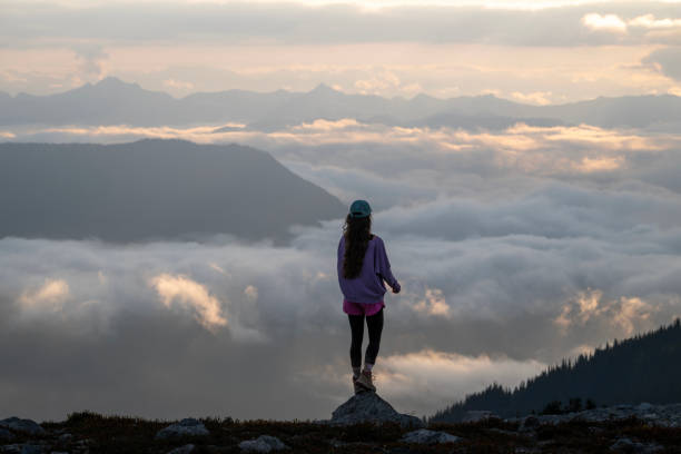 Young woman looks over cloudscape from mountain top at sunrise, Pacific West Coast mountains, British Columbia
