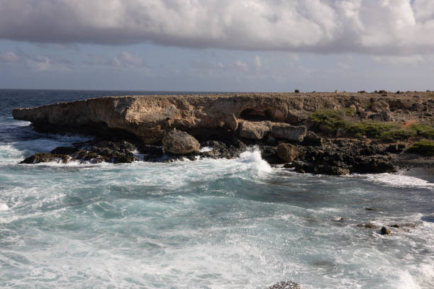 The photograph captures a dramatic Aruba seascape where the powerful waves of the ocean crash against a rugged, rocky shoreline. The foreground showcases the swirling, foamy waters in shades of deep blue and turquoise, contrasting with the dark, jagged rocks. As the waves collide with the shore, white sprays of water erupt into the air, frozen in time by the camera.\nA large, weathered rock formation sits in the middle of the scene, jutting out into the water. Atop this rock, a small stack of stones, known as a cairn, balances precariously, adding a human touch to the otherwise raw and untamed environment. Behind this, a towering rock cliff with a rough, textured surface looms over the crashing waves, its earthy tones contrasting with the deep blues of the sea and sky.\nThe background consists of a dramatic, cloud-filled sky, with a mixture of dark storm clouds and patches of lighter, brighter blue. The combination of the turbulent sea, the rugged rocks, and the moody sky creates a striking image of nature's power and beauty.