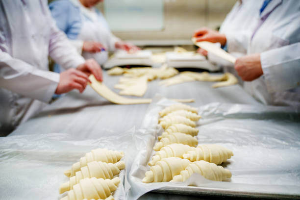 Close-up of multiple bakers working together in a commercial bakery, kneading and shaping fresh dough on a floured stainless steel table. The hands of the workers, dressed in professional bakery uniforms, carefully handle the dough, highlighting teamwork and craftsmanship in bread-making. The scene captures the traditional and industrial baking process, emphasizing food production, artisanal baking, and bakery teamwork. Ideal for concepts related to food industry, baking techniques, and handmade bread preparation.