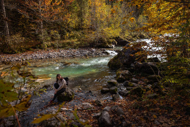 Brunette woman hiker resting on a rock on the shores of a turquoise mountain river in the Austrian Alps during a sunny autumn day