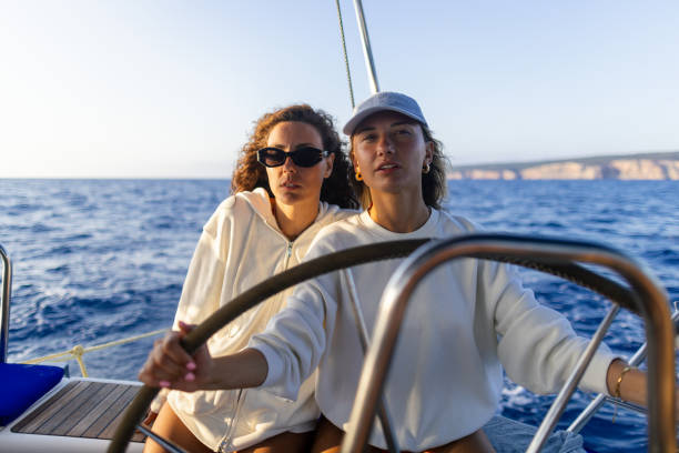 Beautiful woman skipper on a steering wheel navigating a sailboat and showing another woman how to do that.